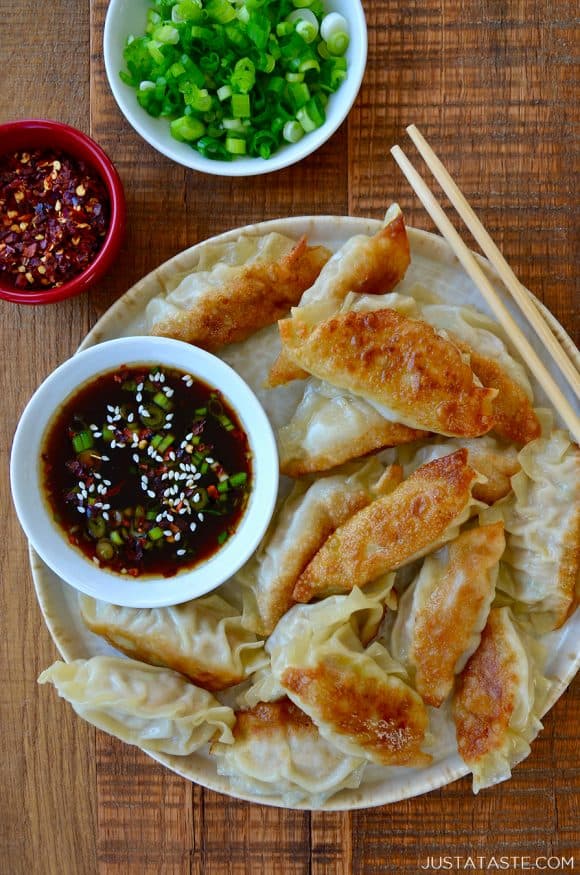 Plate containing easy chicken potstickers next to small bowl with soy dipping sauce and chopsticks next to small bowls with chopped scallions and red pepper flakes