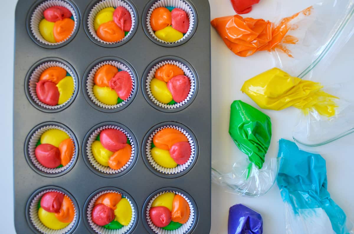 Cupcake wrappers in a muffin tin are filled with red, orange and yellow batter. Nearby, plastic bags are filled with red, orange, yellow, green, blue and purple cupcakes batter, ready to pipe into the tin.