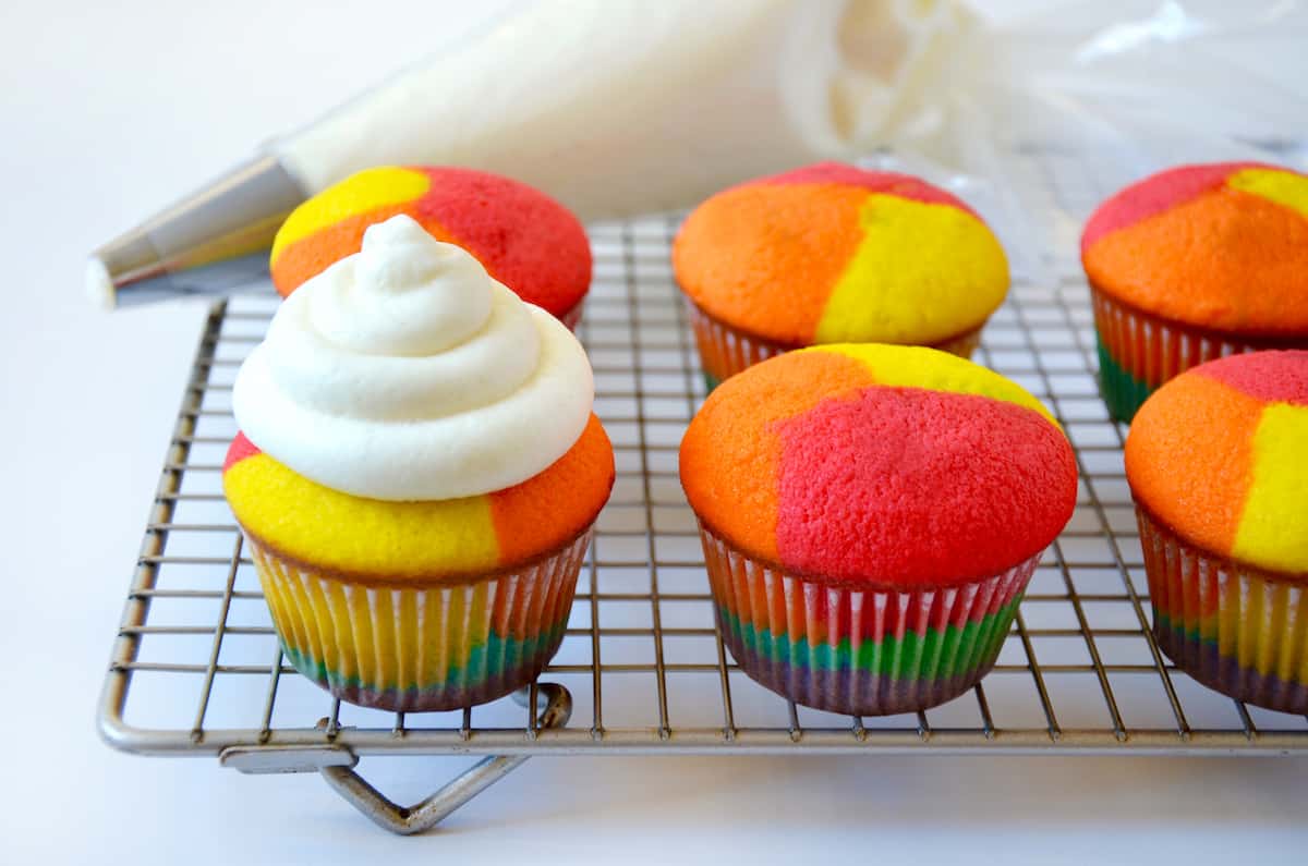 Rainbow cupcakes sit on a baking rack. One is frosted with a dollop of buttercream frosting. In the background is a piping bag filled with frosting and fitted with a pastry tip.