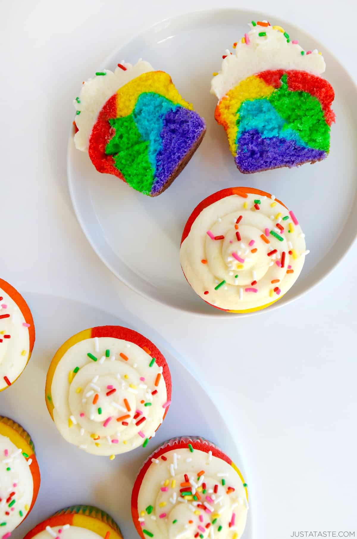 A rainbow cupcake cut in half to show its vibrant center of red, yellow, blue green and purple. More rainbow cupcakes topped with buttercream frosting and rainbow sprinkles are on another plate.