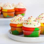 Rainbow cupcakes with white buttercream frosting and rainbow sprinkles are on white plates.