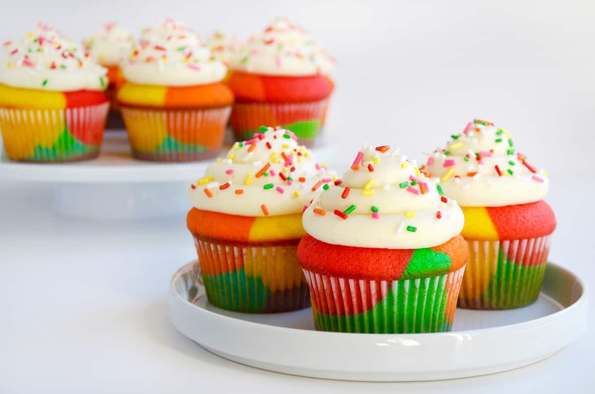 Rainbow cupcakes with white buttercream frosting and rainbow sprinkles are on white plates.