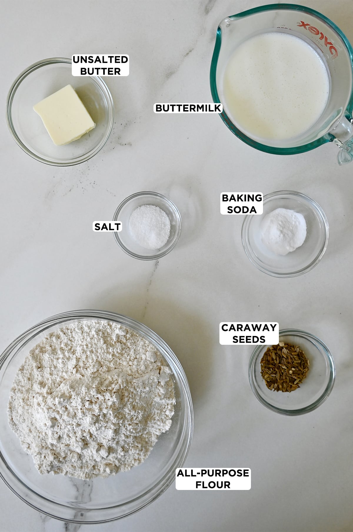 Various sizes of glass bowls containing the ingredients needed to make soda bread, including butter, buttermilk, baking soda, salt, caraway seeds and all-purpose flour.