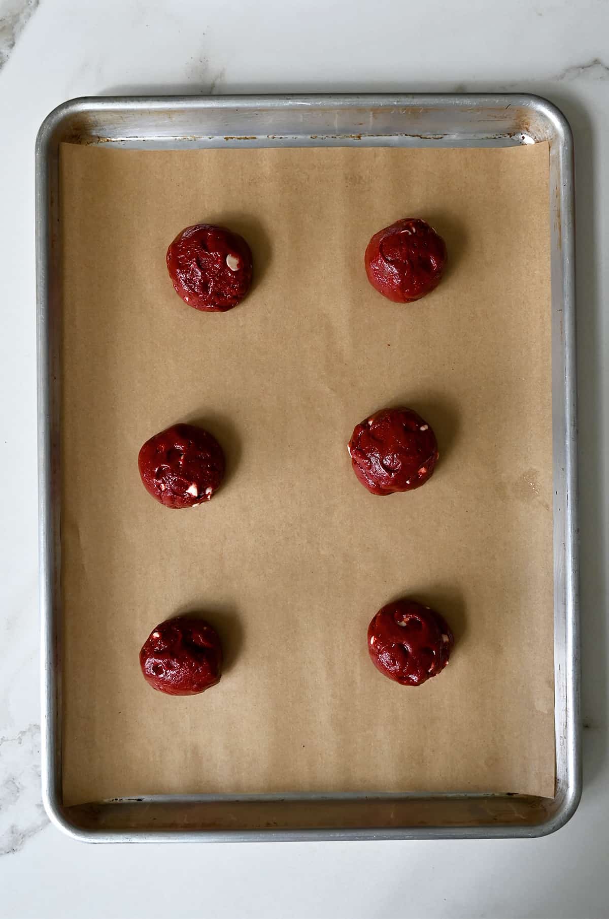 Six balls of red velvet cookie dough on a parchment paper-lined baking sheet.
