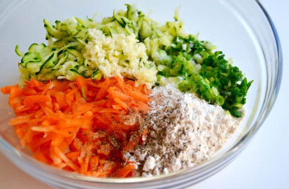 Glass bowl containing shredded zucchini, minced garlic, sliced scallions, flour, spices and shredded carrots