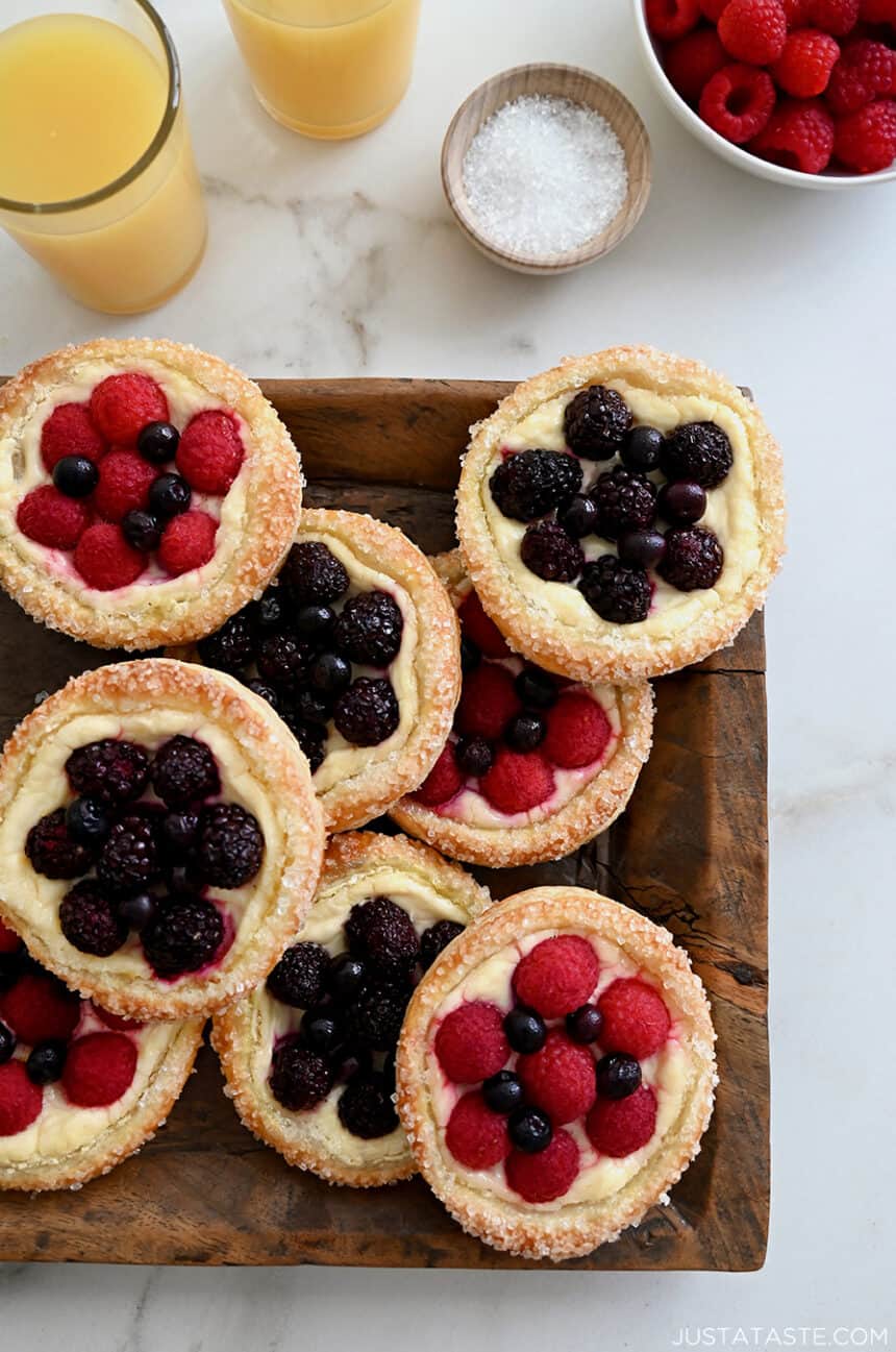 A top-down view of Fruit and Cream Cheese Breakfast Pastries on a wood serving platter next to glasses filled with orange juice and a bowl with fresh raspberries