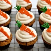 Carrot cake cupcakes with swirled cream cheese frosting and candy melt toppers on a cooling rack.