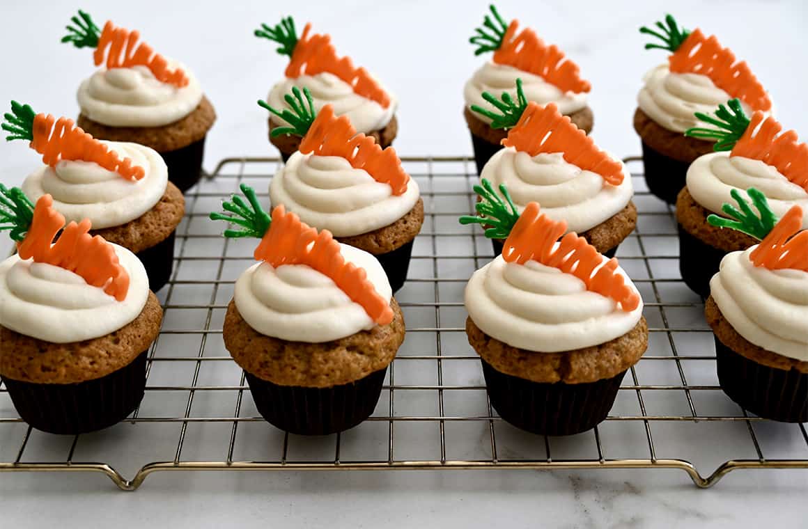 Carrot cupcakes with silky frosting and carrot candy melt toppers atop a wire cooling rack.