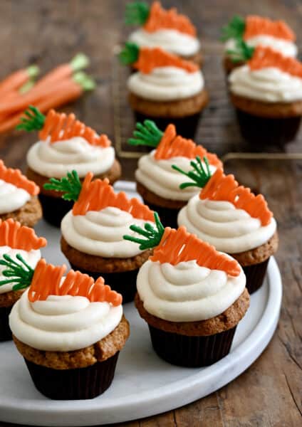 Carrot cupcakes topped with piped cream cheese frosting and carrot candy melt toppers on a white plate.