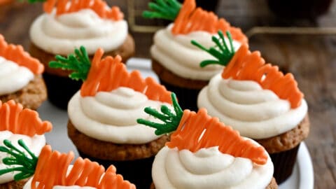 Carrot cupcakes topped with piped cream cheese frosting and carrot candy melt toppers on a white plate.