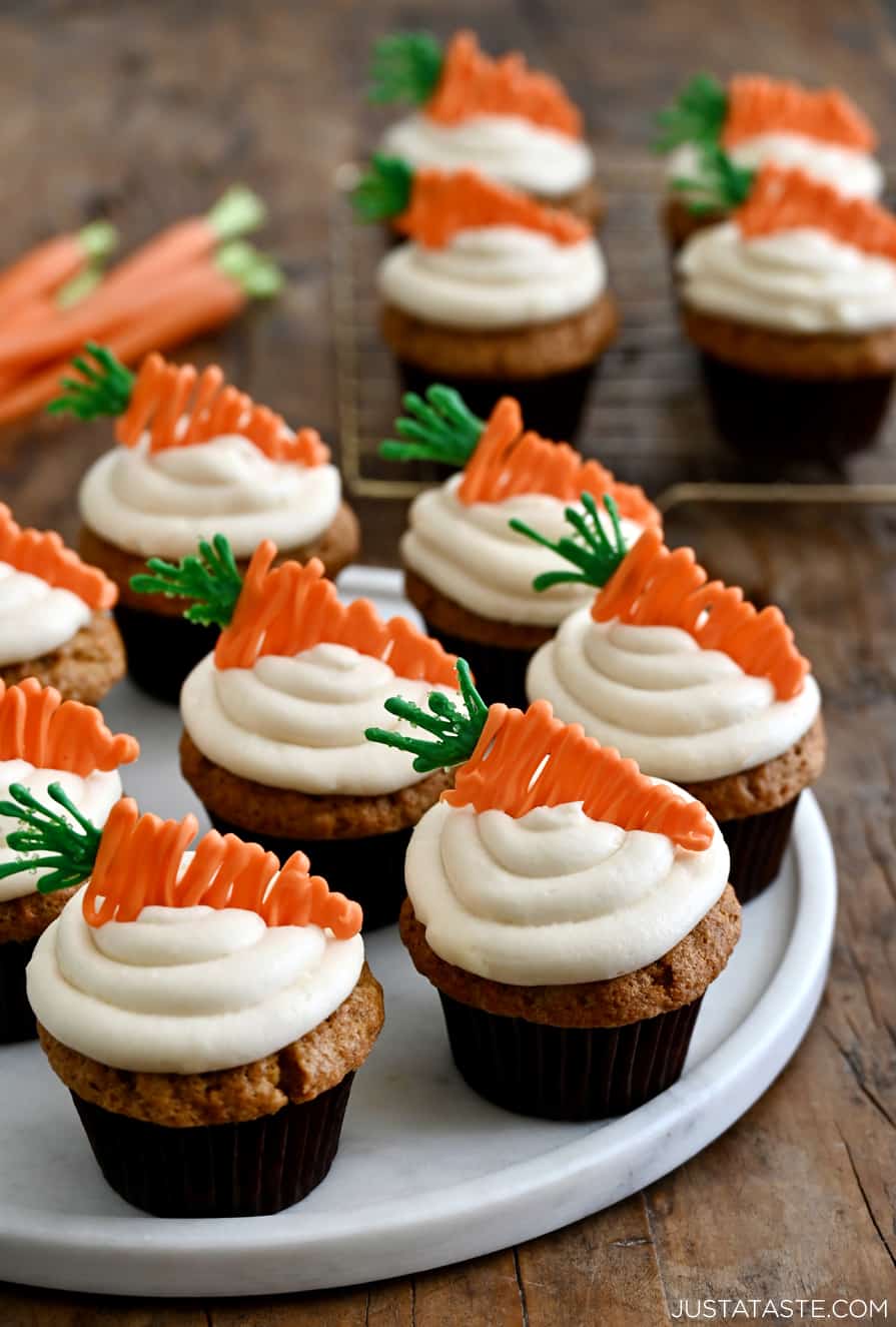 Carrot cupcakes topped with piped cream cheese frosting and carrot candy melt toppers on a white plate.