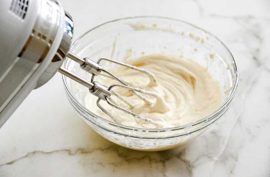 A handheld mixer resting against a clear bowl containing whipped cream cheese