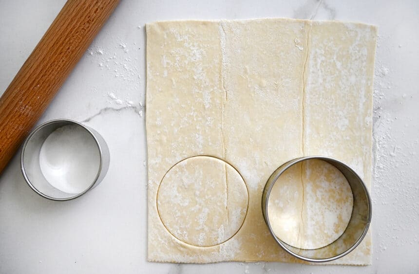 A circular cookie cutter atop a sheet of puff pastry next to a rolling pin