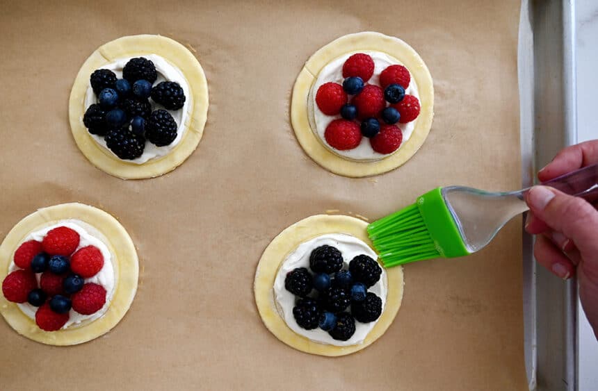 A hand with a pastry brush applies an egg wash to unbaked pastries on a parchment paper-lined baking sheet