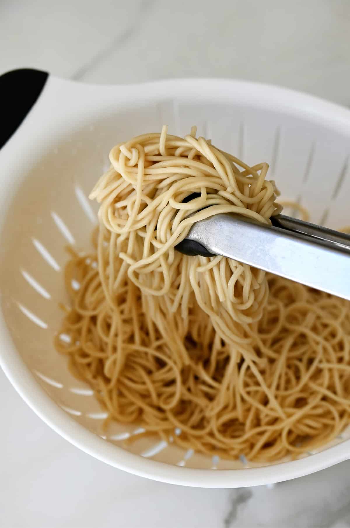 Tongs grabbing udon noodles in a strainer.