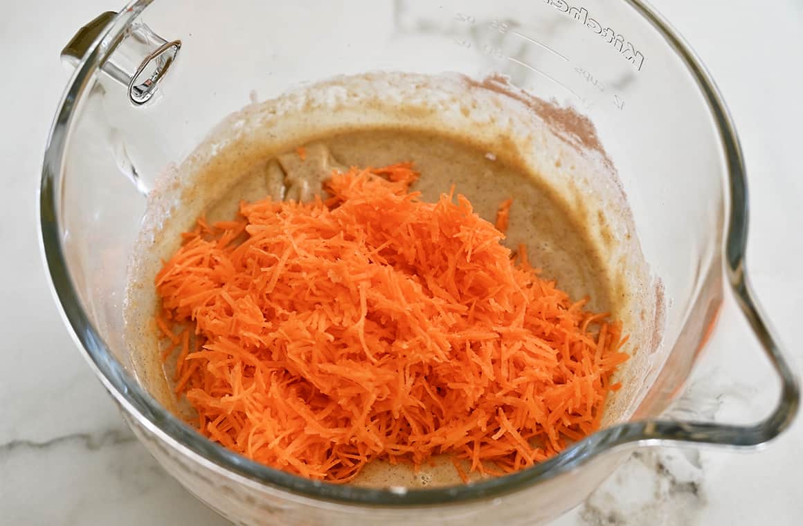 A large clear bowl containing shredded carrots over cake batter.