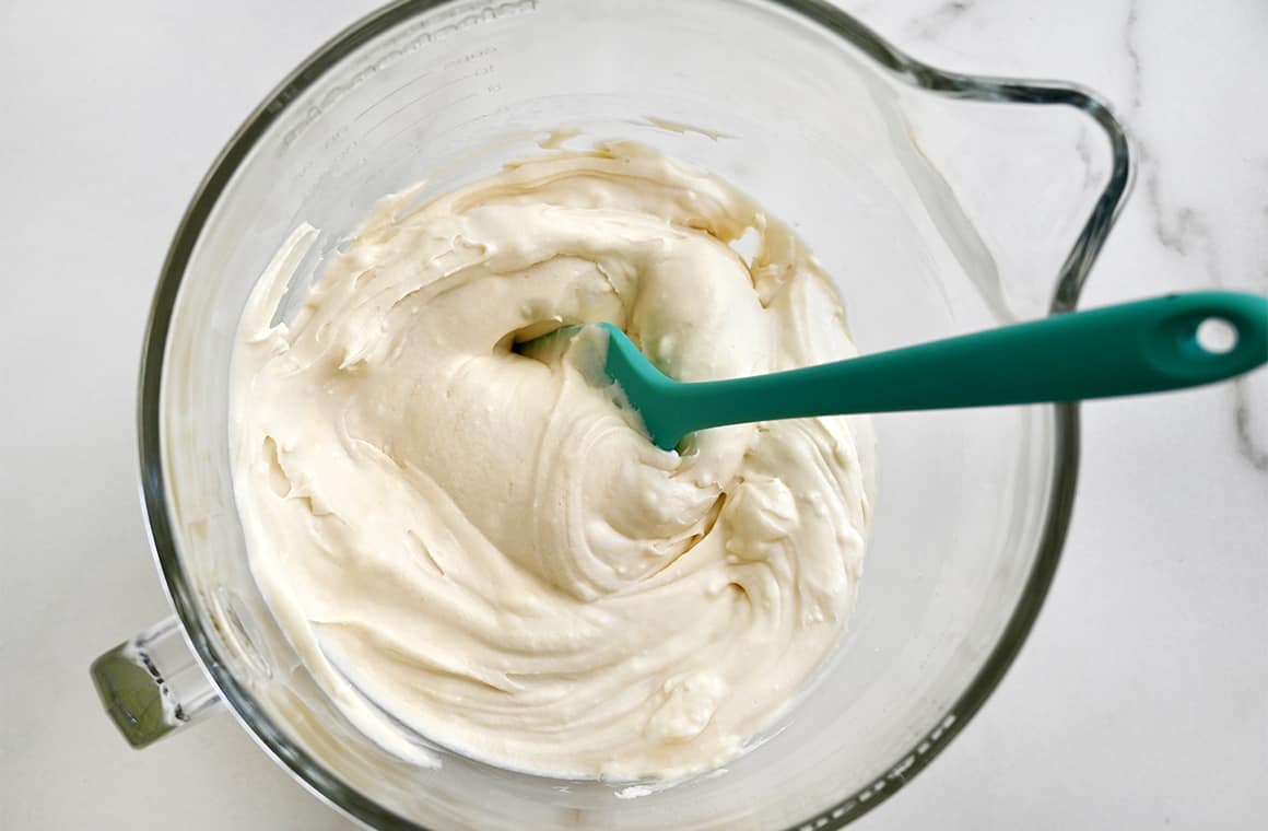 A top-down view of a clear bowl containing cream cheese frosting and a teal spatula.