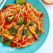 Top-down view of a blue plate containing Teriyaki Chicken Stir-Fry over noodles next to two small ramekins filled with crushed red pepper flakes and sesame seeds.