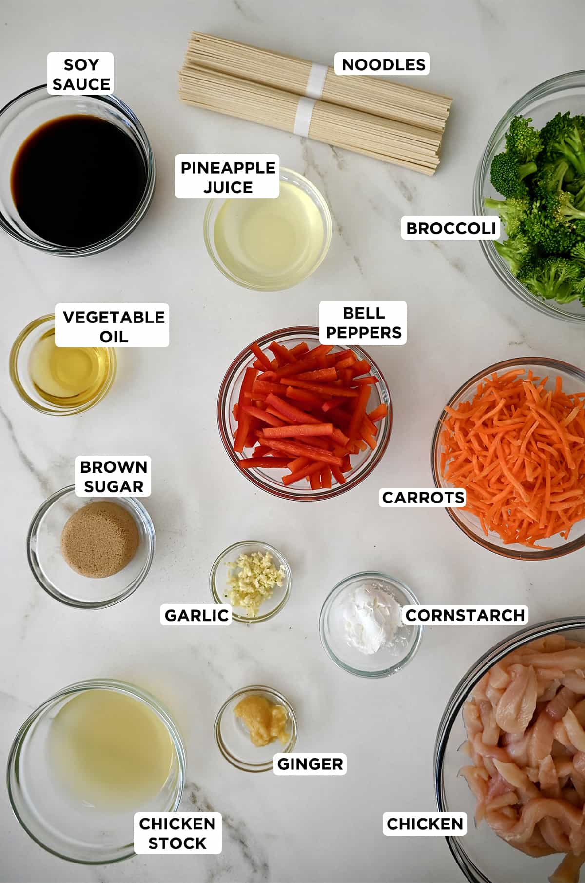 Various sizes of glass bowls containing ingredients for stir fry teriyaki chicken, including soy sauce, pineapple juice, broccoli, bell peppers, shredded carrots, cornstarch, minced garlic, brown sugar, vegetable oil, minced ginger, chicken stock, chicken breast slices and udon noodles. 