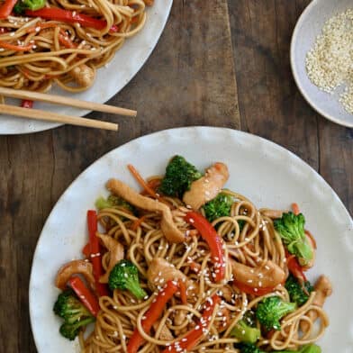 Two dinner plates containing Teriyaki Chicken Stir-Fry over noodles next to a small bowl filled with sesame seeds and a small bowl containing sambal oelek.