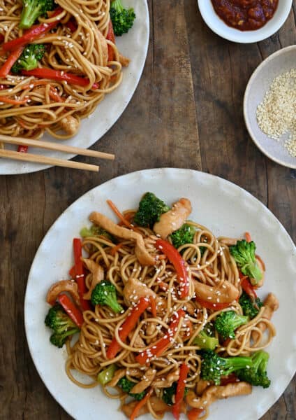 Two dinner plates containing Teriyaki Chicken Stir-Fry over noodles next to a small bowl filled with sesame seeds and a small bowl containing sambal oelek.