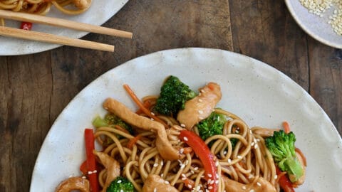 Two dinner plates containing Teriyaki Chicken Stir-Fry over noodles next to a small bowl filled with sesame seeds and a small bowl containing sambal oelek.