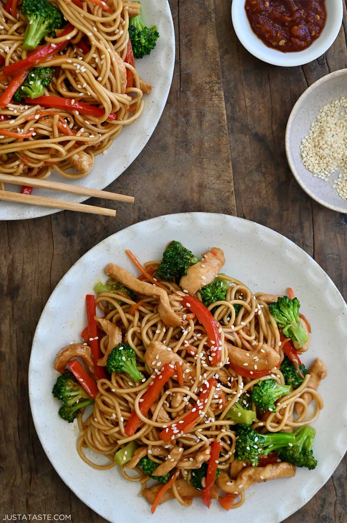 Two dinner plates containing Teriyaki Chicken Stir-Fry over noodles next to a small bowl filled with sesame seeds and a small bowl containing sambal oelek.