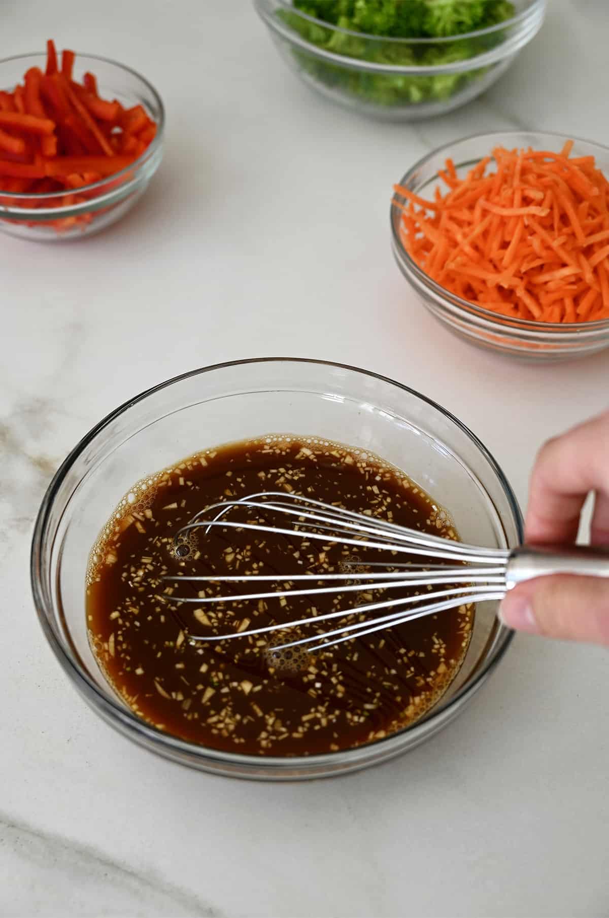 A hand holds a whisk into a small glass bowl containing teriyaki stir fry sauce.