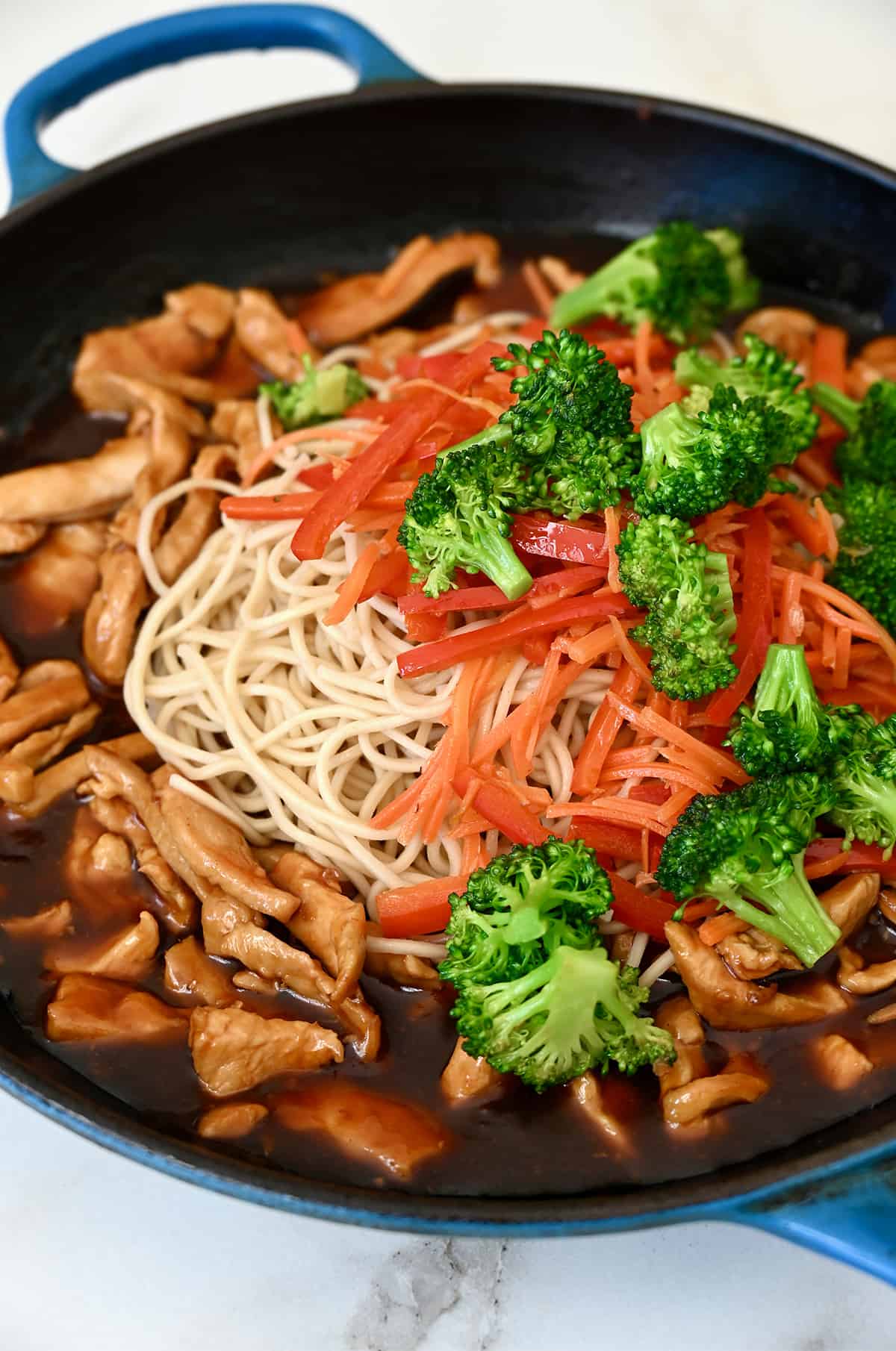 A large skillet containing teriyaki sauce, cooked chicken, noodles and stir-fried veggies.