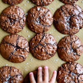 A child's hand reaches for a flourless cookie with chocolate chips topped with large-flake sea salt