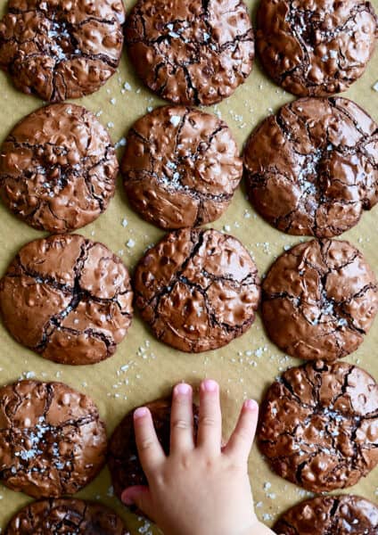 A child's hand reaches for a flourless cookie with chocolate chips topped with large-flake sea salt