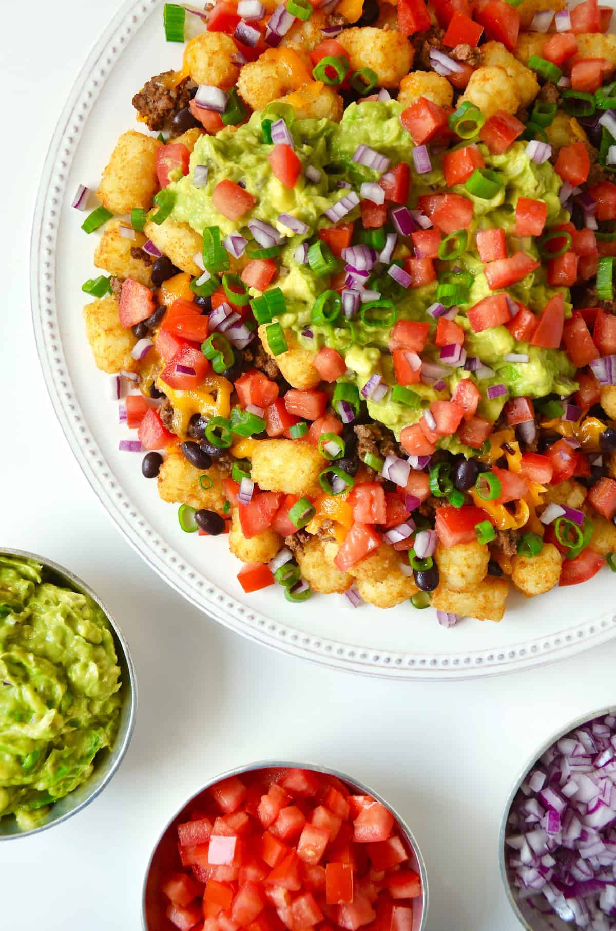 A white serving platter with loaded totchos (tater tot nachos) topped with diced tomatoes, scallions, black beans and diced avocado.