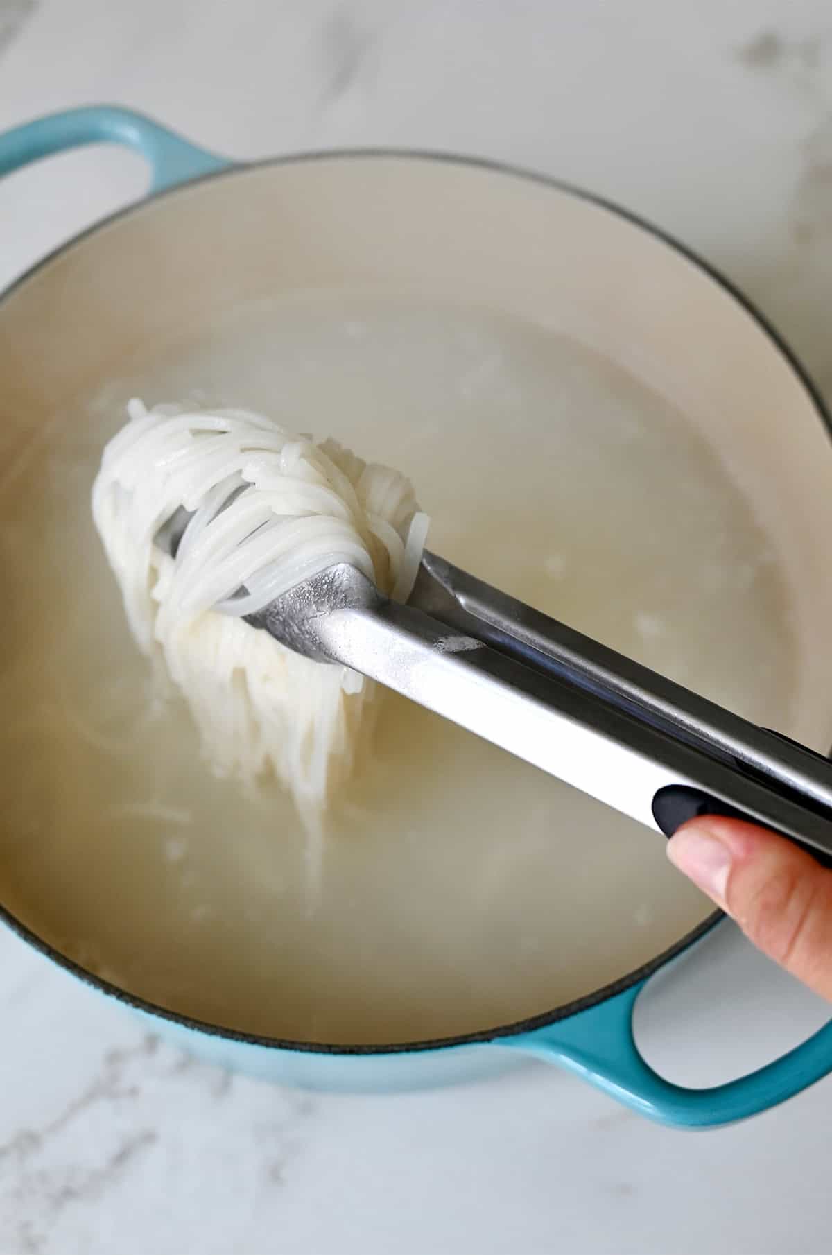 A hand holding tongs pulls cooked rice noodles out of water in a large stockpot.