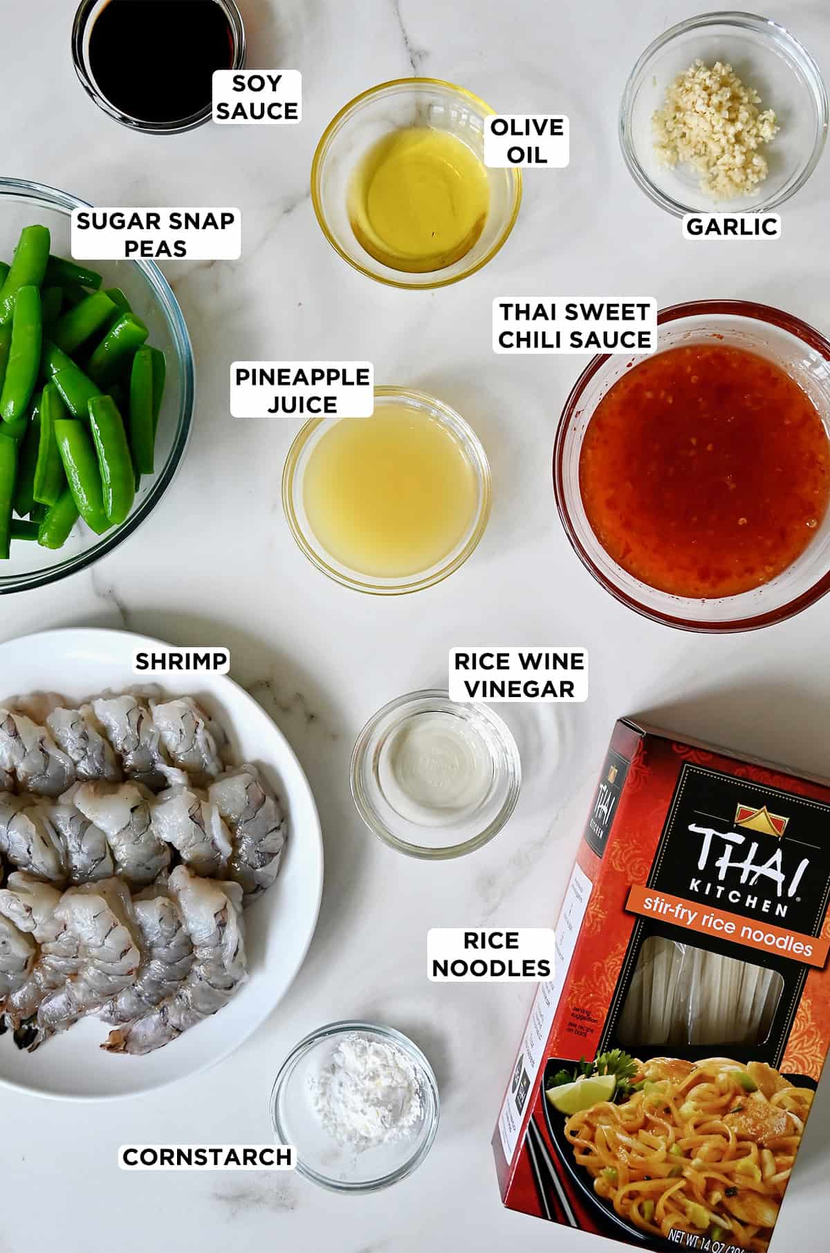 A box of Thai Kitchen rice noodles next to various sizes of glass bowls filled with cornstarch, raw shrimp, rice wine vinegar, pineapple juice, sugar snap peas, olive oil, diced garlic and Thai sweet chili sauce. 