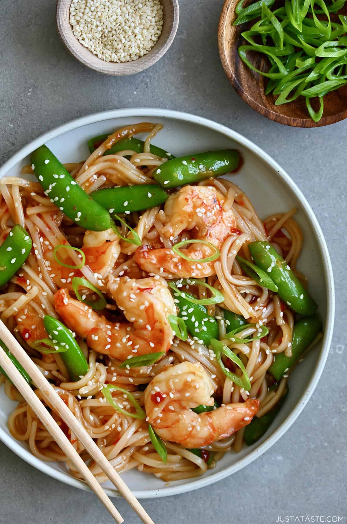 Stir-fried sweet and sour shrimp with snap peas garnished with sesame seeds atop rice noodles in a white bowl with chopsticks.