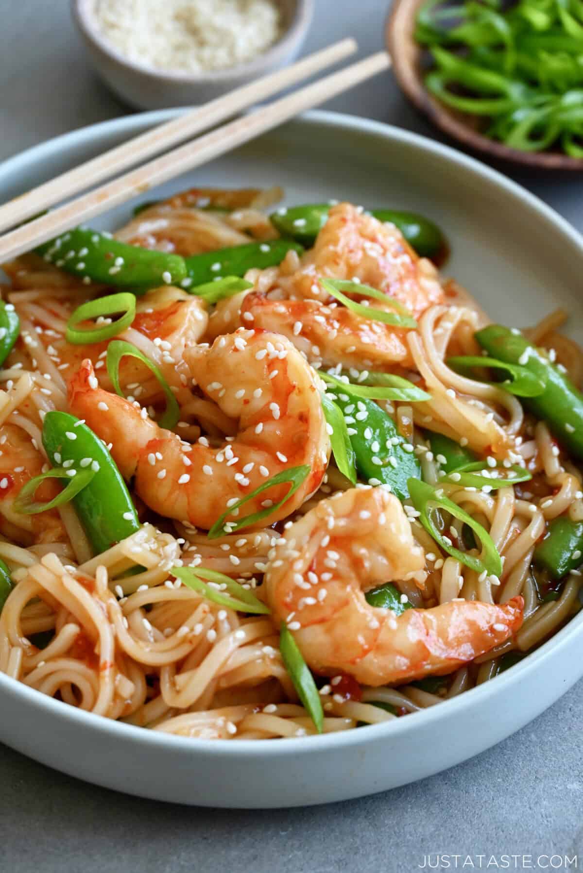 Sweet and sour shrimp stir-fry with rice noodles garnished with sesame seeds in a shallow bowl with chopsticks.