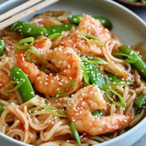 Sweet and sour shrimp stir-fry with rice noodles garnished with sesame seeds in a shallow bowl with chopsticks.