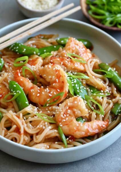 Sweet and sour shrimp stir-fry with rice noodles garnished with sesame seeds in a shallow bowl with chopsticks.