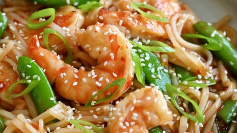 Sweet and sour shrimp stir-fry with rice noodles garnished with sesame seeds in a shallow bowl with chopsticks.