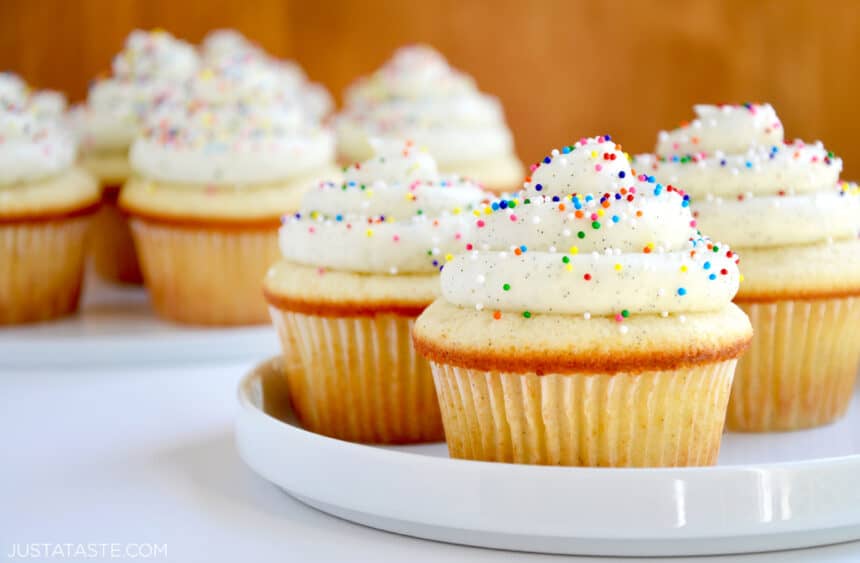 White serving plate containing Vanilla Bean Cupcakes with Vanilla Buttercream Frosting and rainbow sprinkles with more cupcakes in blurred out in the background