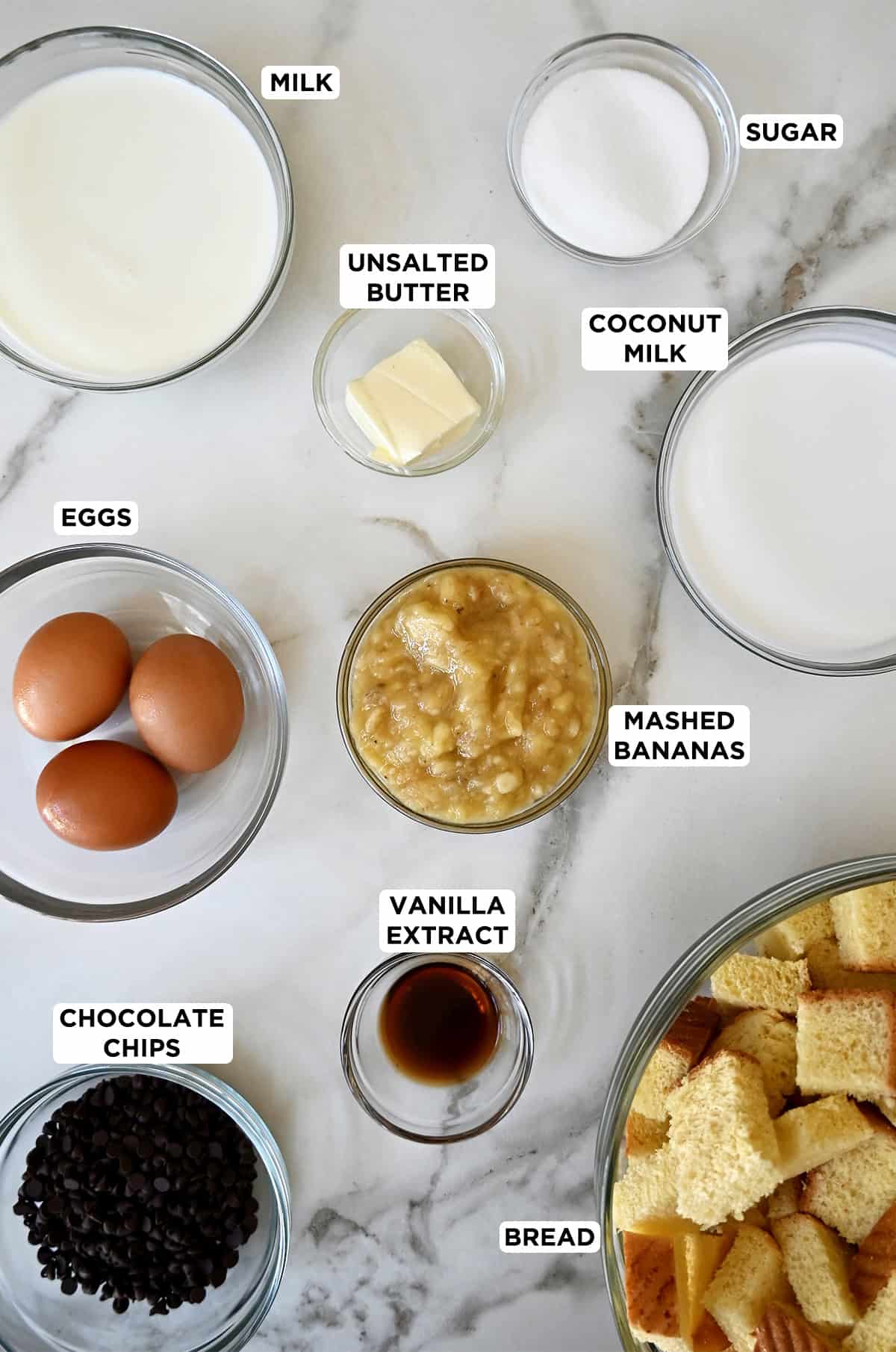 Various sizes of glass bowls containing coconut milk, eggs, chocolate chips, mashed bananas, vanilla extract, milk, cubed brioche bread and butter.