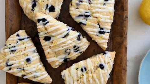 A top-down view of Glazed Lemon Blueberry Scones on a wood serving platter next to a bowl filled with fresh blueberries and a lemon cut in half.