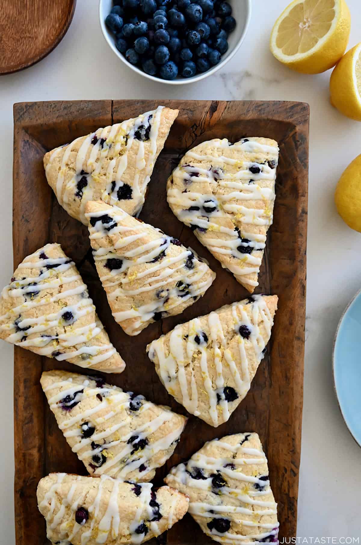 A top-down view of Glazed Lemon Blueberry Scones on a wood serving platter next to a bowl filled with fresh blueberries and a lemon cut in half.