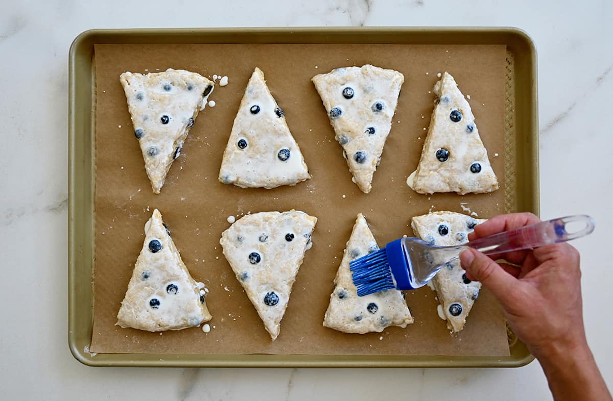 A hand holding a rubber brush apples heavy cream to the top of unbaked scones on a parchment paper-lined baking sheet.