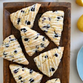 A top-down view of Glazed Lemon Blueberry Scones on a wood serving platter next to a bowl filled with fresh blueberries and a lemon cut in half