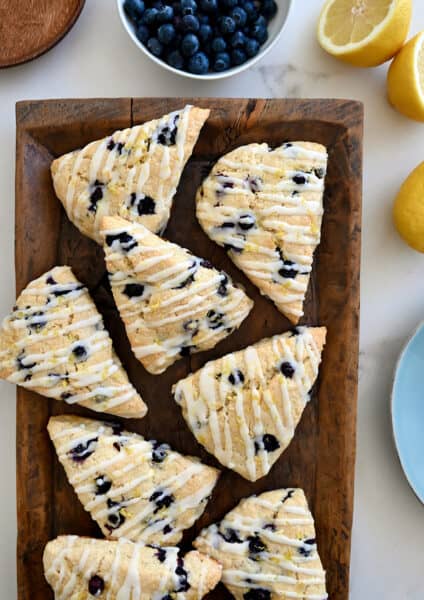 A top-down view of Glazed Lemon Blueberry Scones on a wood serving platter next to a bowl filled with fresh blueberries and a lemon cut in half
