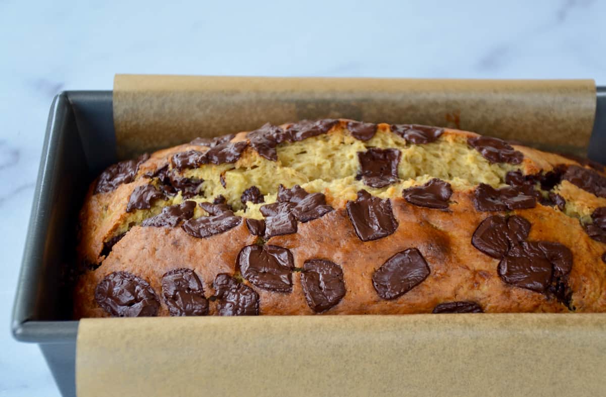 A loaf of banana bread studded with chocolate chunks in a bread pan lined with parchment paper.