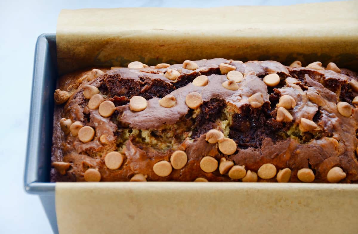 A loaf of banana bread studded with peanut butter chips in a bread pan lined with parchment paper.