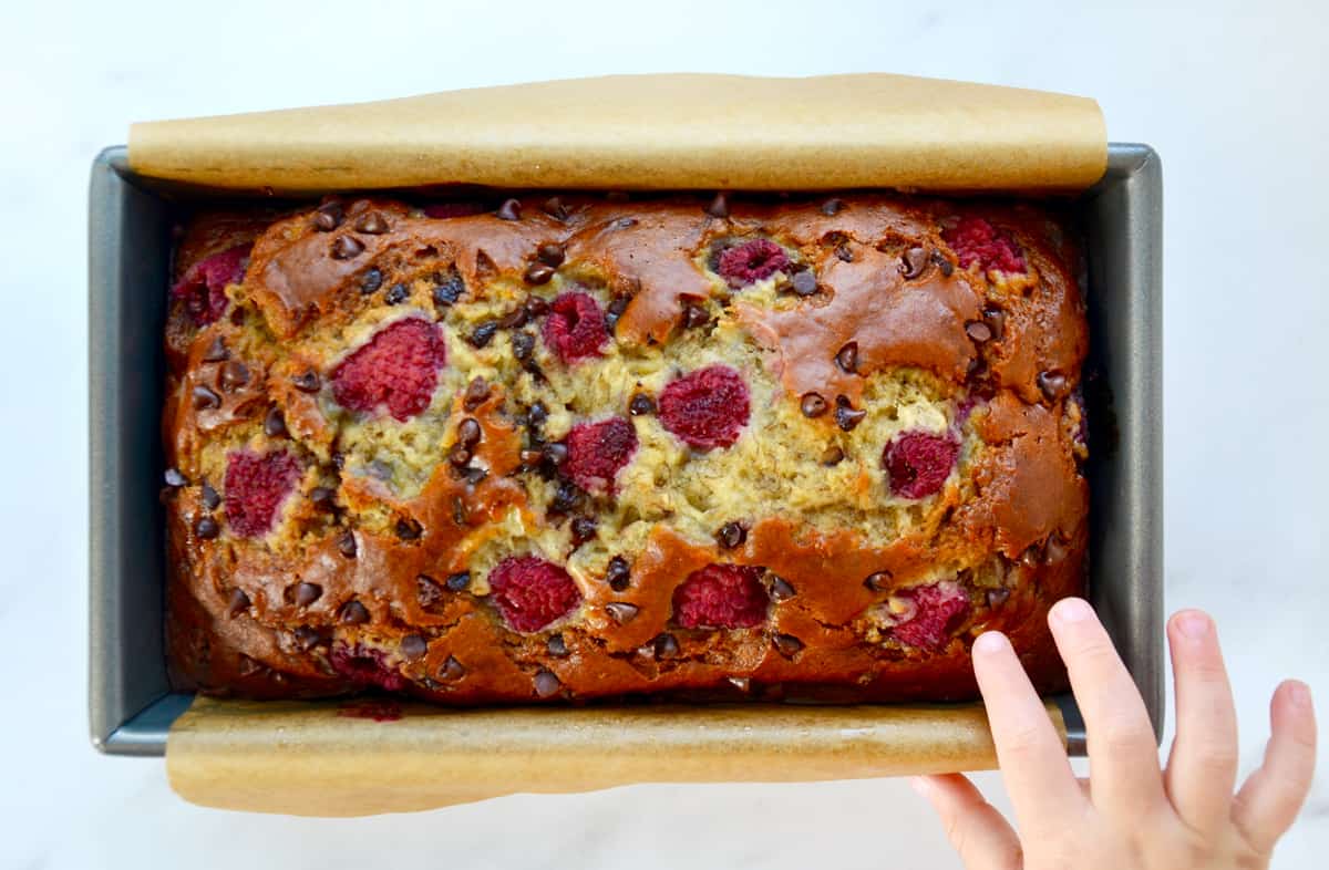 A child's hand reaches for a loaf of raspberry chocolate chip banana bread in a loaf pan.