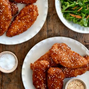 Orange chicken tenders on a plate garnished with sesame seeds next to a plate topped with mixed salad greens.